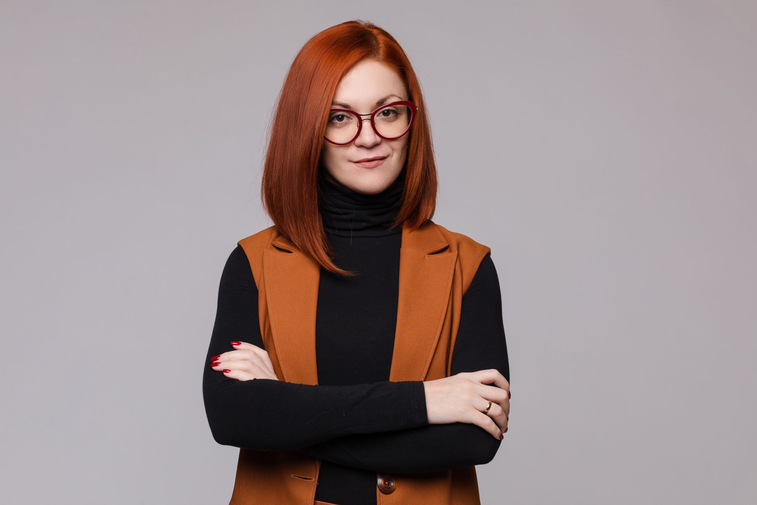 Portrait of smiling red-haired business woman in glasses posing looking at camera medium shot. Beauty European female boss standing with crossed hand isolated at white studio background